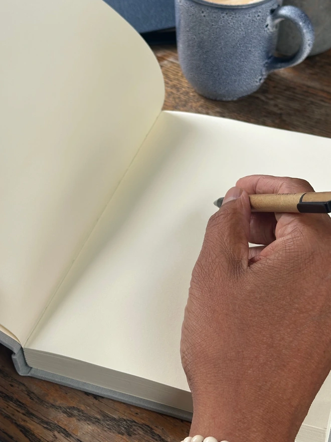 a really close up image of someone poised to write in a journal. The journal is open flat on the desk and it has plain pages.