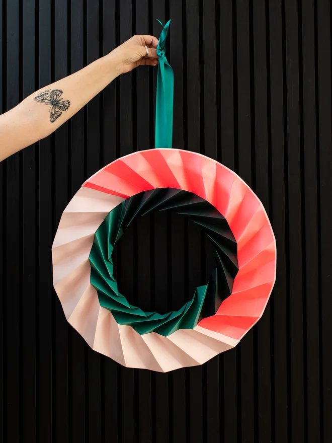 Woman's arm stretched out with a modern origami wreath hanging in neon pink, pale pink, dark green and jade greeen.