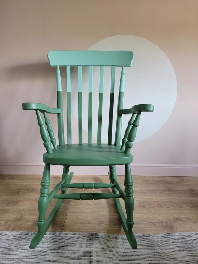 Green and mint painted wooden rocking chair front on with a paink wall and rug on wooden floor