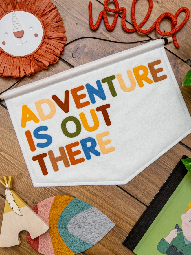 White felt banner with the words "Adventure is out there" written on it in a mix a earthy, jungle themed colours