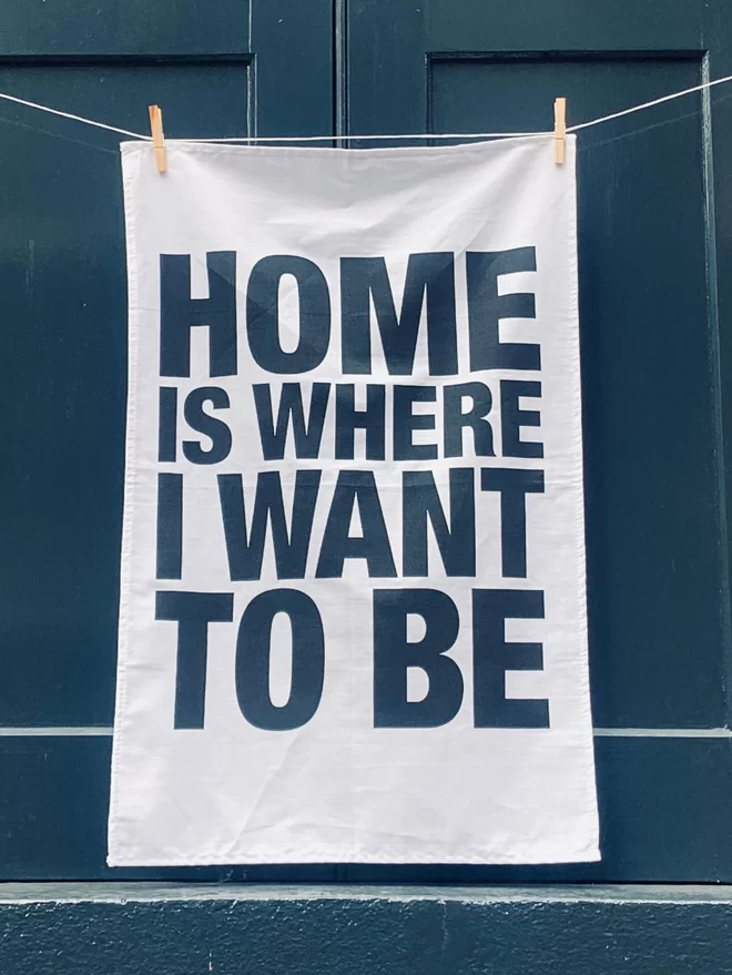 London Drying Home Is Where I Want To Be black screen printed text on white tea towel hanging washing line style in front of dark grey window shutter