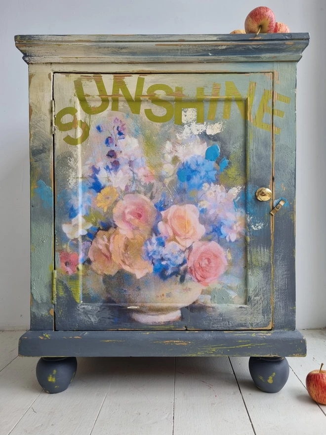 Image shows a painted cupboard in grey with a floral decoupage centrepiece. There is text on the piece in the contrasting green of the undercoat that says ‘sunshine’. The flowers are pink, peach, blue and cream. The cupboard is solid pine with chunky bun feet