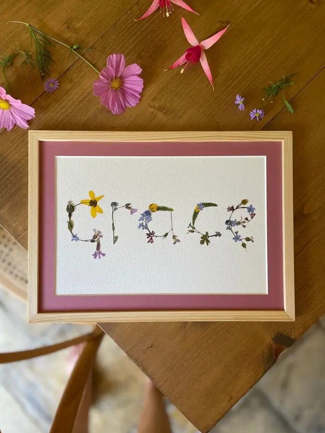 Personalised Original Pressed Flower Name In Frame, a framed flower name prints sitting on a wooden surface. 