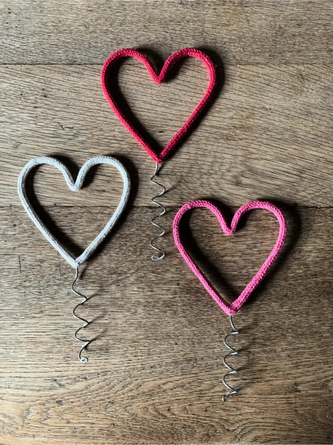 A red, a white and a pink string wrapped wire heart Christmas tree topper on an oak table