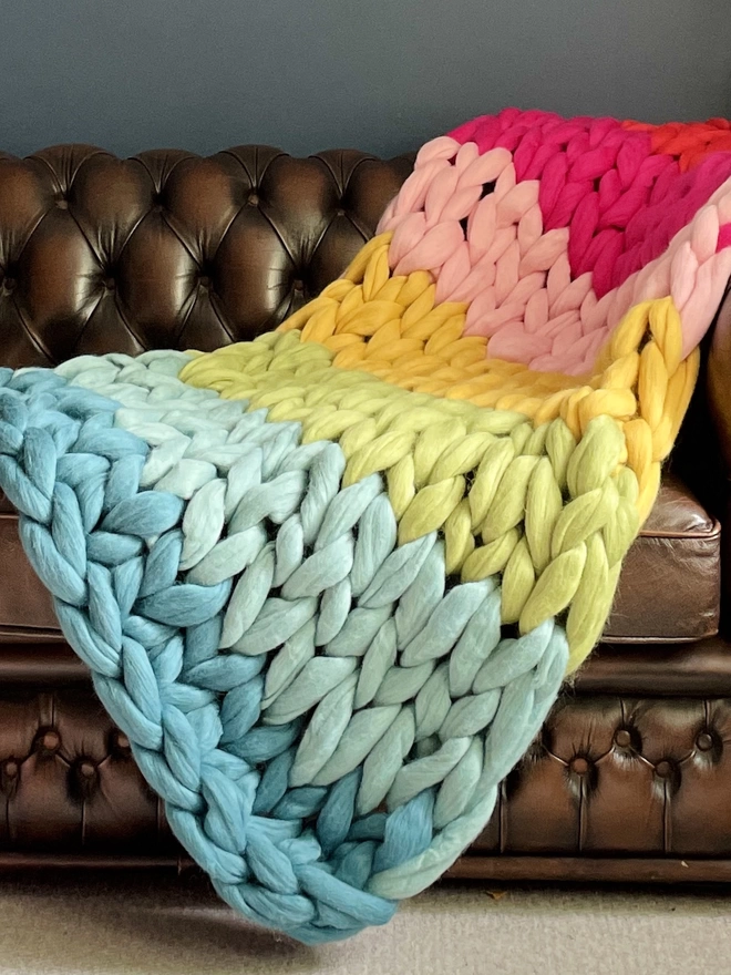 A close up of A rainbow merino arm knitted blanket cascading down the side of a brown leather Chesterfield sofa, before a dark blue wall