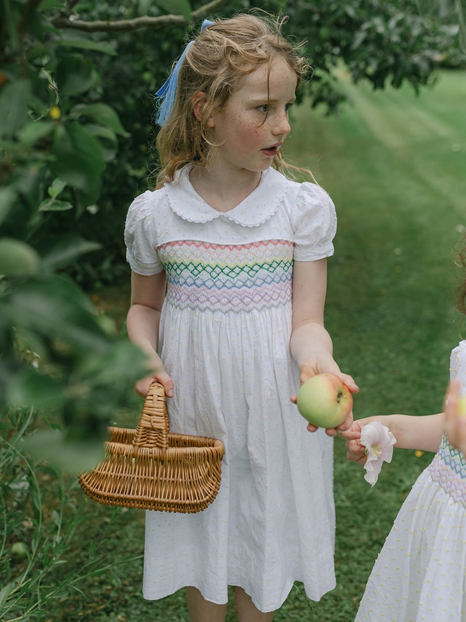 A girl holds a basket wearing a white dress with rainbow smocking and a peter pan collar