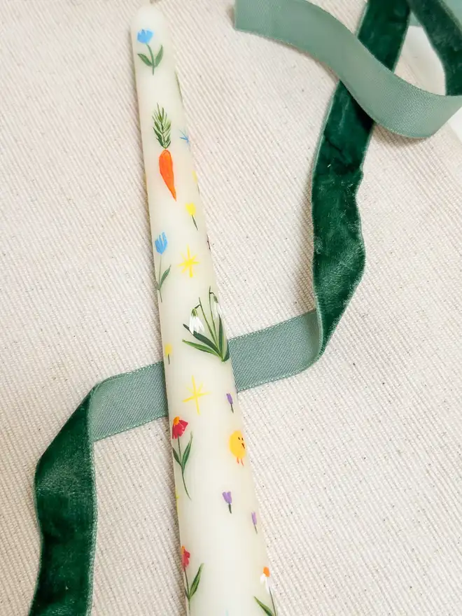 Spring Fun Candle, a white spring candle lying on a plain surface accompanied by a green ribbon. It's floral design can be seen wrapping around it. 