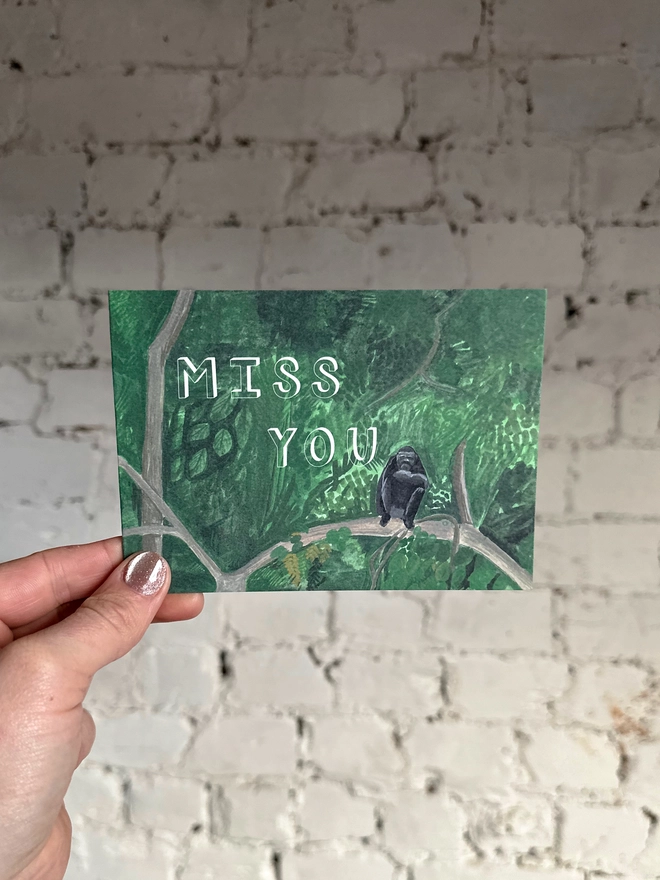 a greetings card featuring an illustration of a chimp sitting in a jungle landscape with the text “miss you”