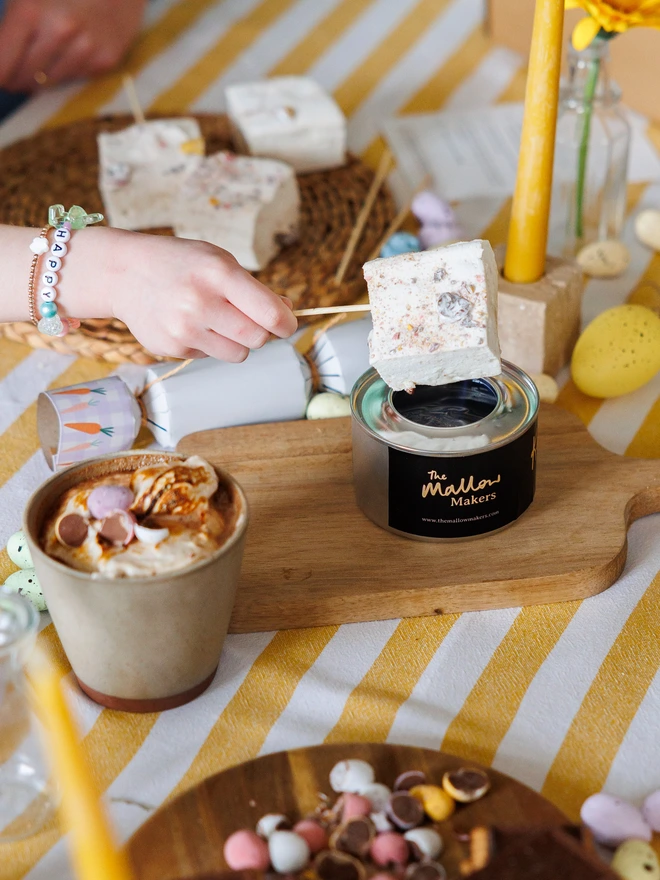 The Mallow Makers Easter Luxury Smores and Hot Chocolate Kit with child's hand holding a jumbo mini egg marshmallow over a mini gel burner on a striped yellow table cloth beside a mug of hot chocolate