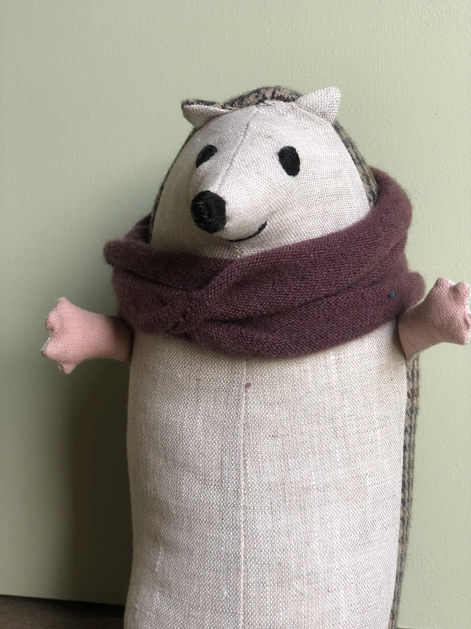 Close up of handmade toy textile hedgehog, made from natural linen (front) and brown tweed (back). Wearing a burgundy woollen scarf, with embroidered eyes, nose and mouth