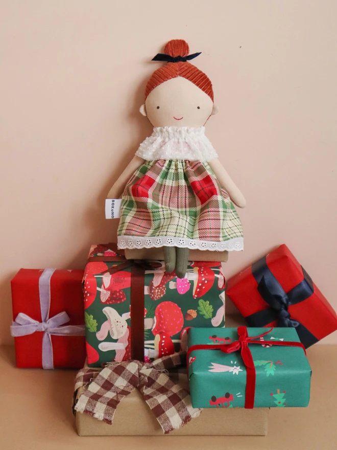 Handmade cloth doll with red brown corduroy hair in a top knot tied with a navy velvet bow, wearing a white textured ruffle collar, a red and green tartan dress with lace trim, and dark green tights.
