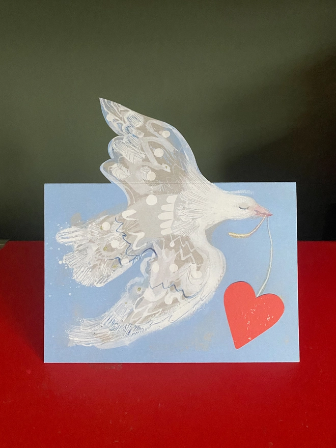 Esther Kent greetings card showing white flying dove in a light blue background, carrying a vibrant red heart on a string