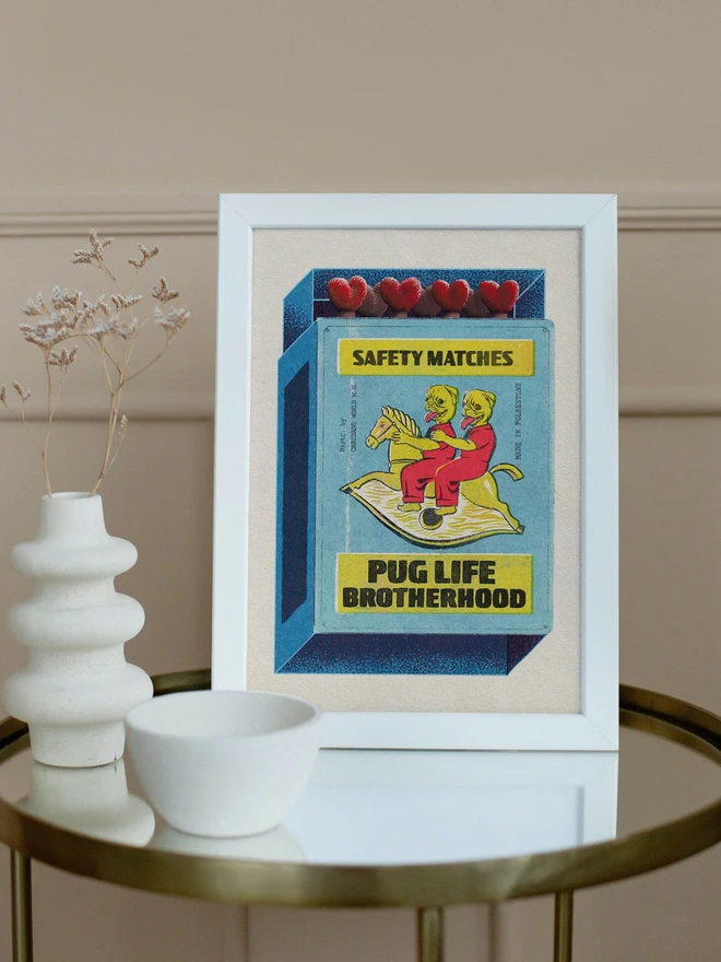 A light blue matchbox with a yellow label that reads "SAFETY MATCHES" at the top and "HAPPY BIRTHDAY" at the bottom. The matchbox is partially open, revealing five red heart-shaped match heads. The main illustration on the matchbox features two yellow, dog-like figures with red outfits and long red tongues, riding a single yellow rocking horse. Text on the side of the illustration reads "Distr. by Caridade World M.G." and "MADE IN THE U.K."