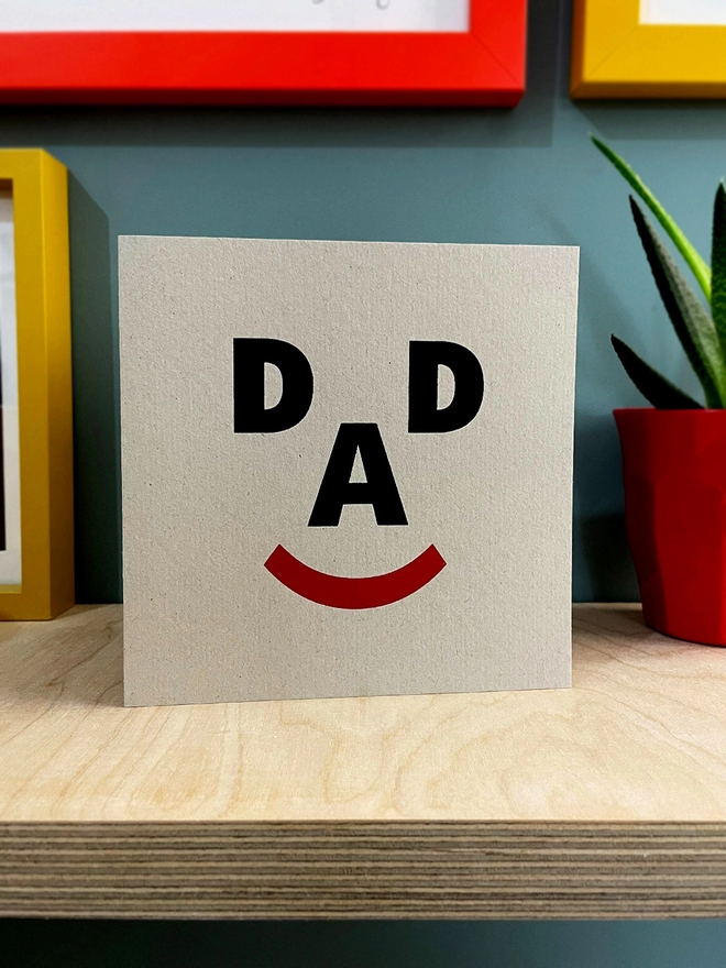 The letters D, A, D and a closed bracket symbol make up a happy face, screenprinted on recycled grey card. Stood on a plywood shelf with hint of frames around, a plant to one side and a duck egg blue wall.