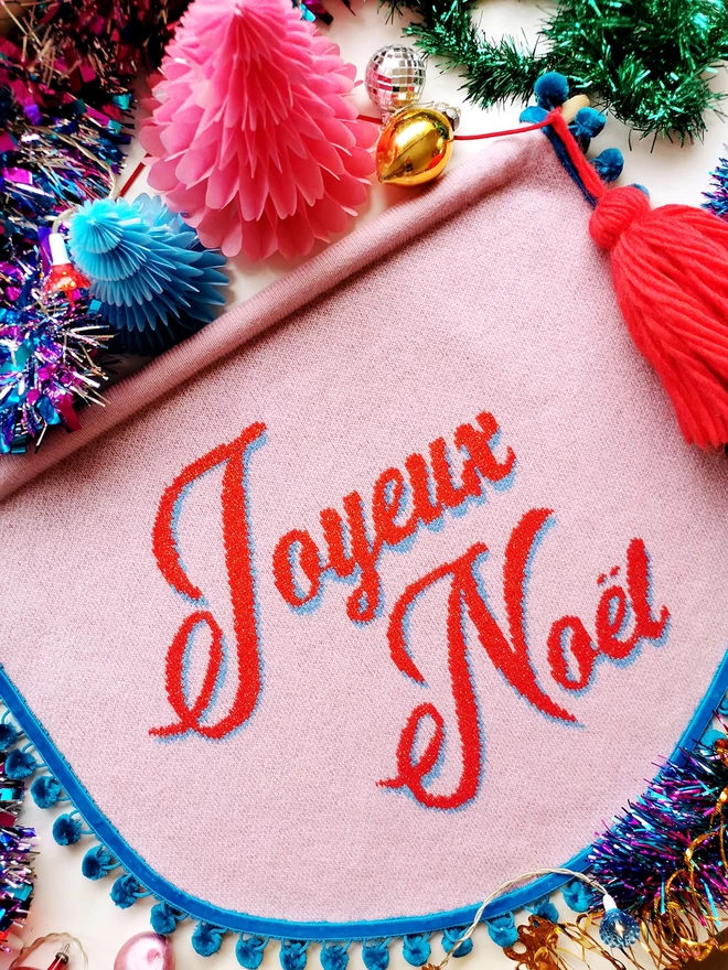 Image shows a table of brightly coloured retro Christmas decorations. Amongst the tinsel, coloured lights, glass baubles and paper christmas trees is a pink knitted scallop shaped wall hanging that reads Joyeux Noel in bright red sparkly letters.