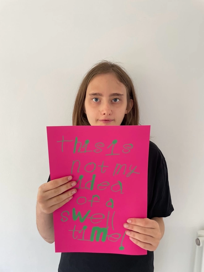 Piper holding up a copy of her digital print green handwritten font on a bright pink back ground