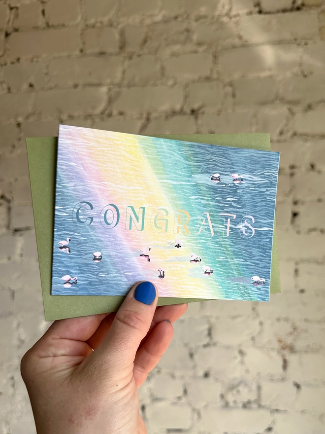 a greetings card featuring an illustration of of flamingos in a lake with a rainbow reflection with the text “CONGRATS” with a green envelope