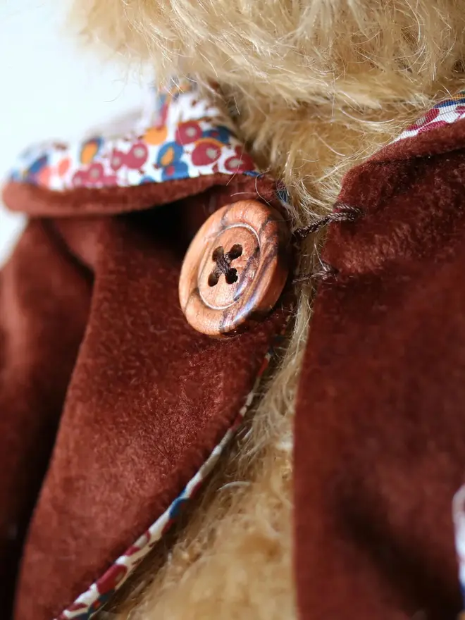 Brown teddy bear wearing a burgundy cloak with flower details and a brown button, sitting upright against a white background.