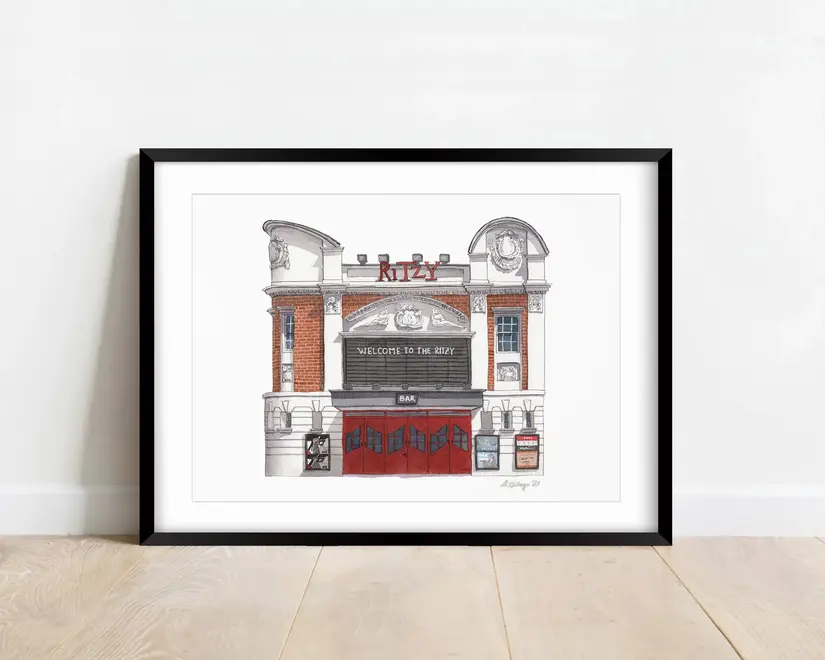 Brixton Ritzy Cinema Giclée Print (Unframed), a print of a cinema sitting in a black frame on a wooden surface against a white backdrop. 