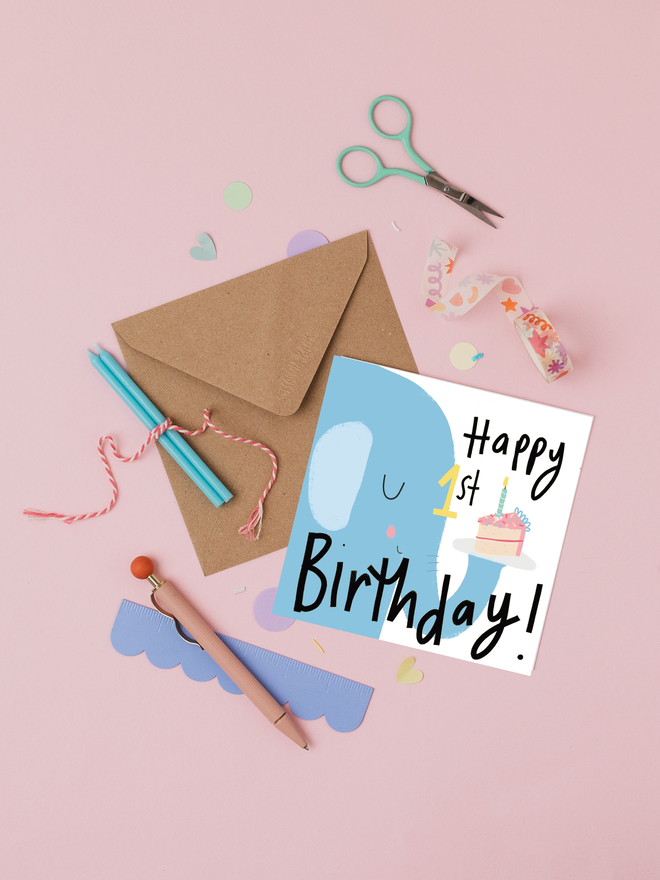 'happy 1st birthday' illustrated elephant card. card featuring a blue elephant holding a cake on its trunk. 