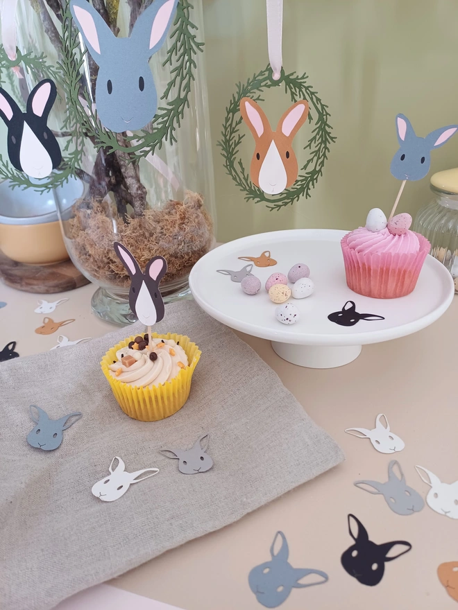 Easter bunny cupcake toppers in iced cupcakes on table decorated for Easter. In background, Easter Tree decorated with Easter Bunny Decorations