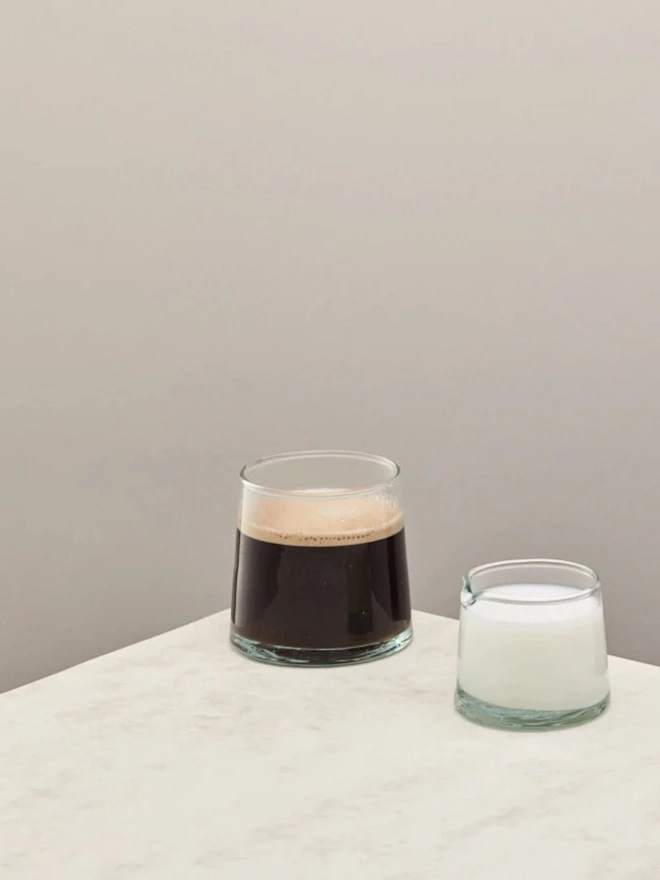 Low Jug, a small glass jug sitting on a counter top against a plain backdrop. It is filled by milk and accompanied by another glass. 