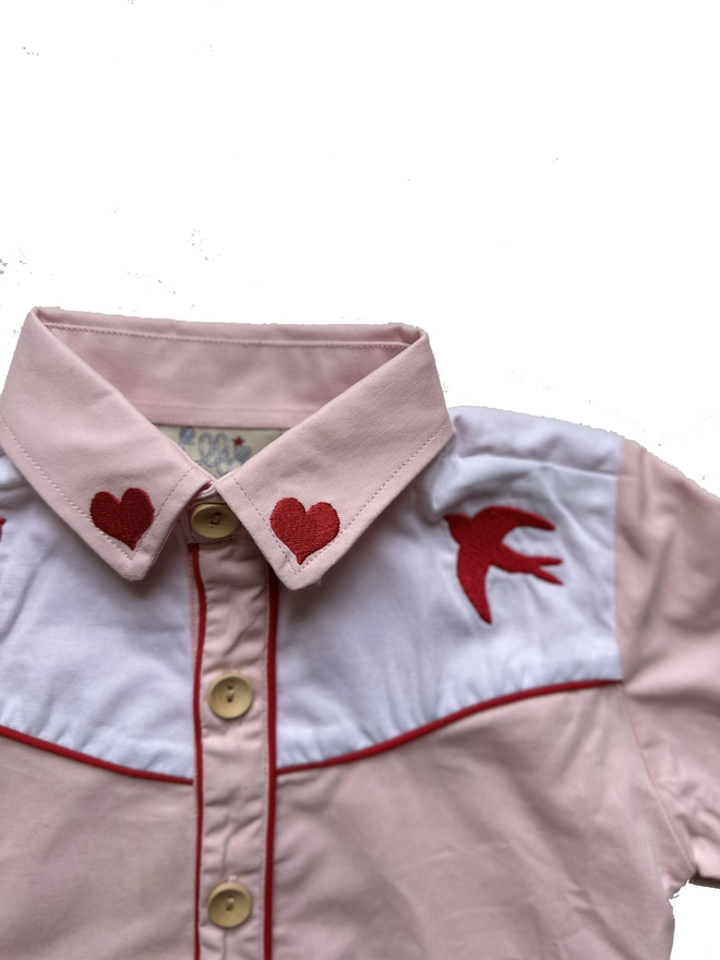 A detail of a pink and white cowgirl shirt with red piping and heart and swallow embroidery