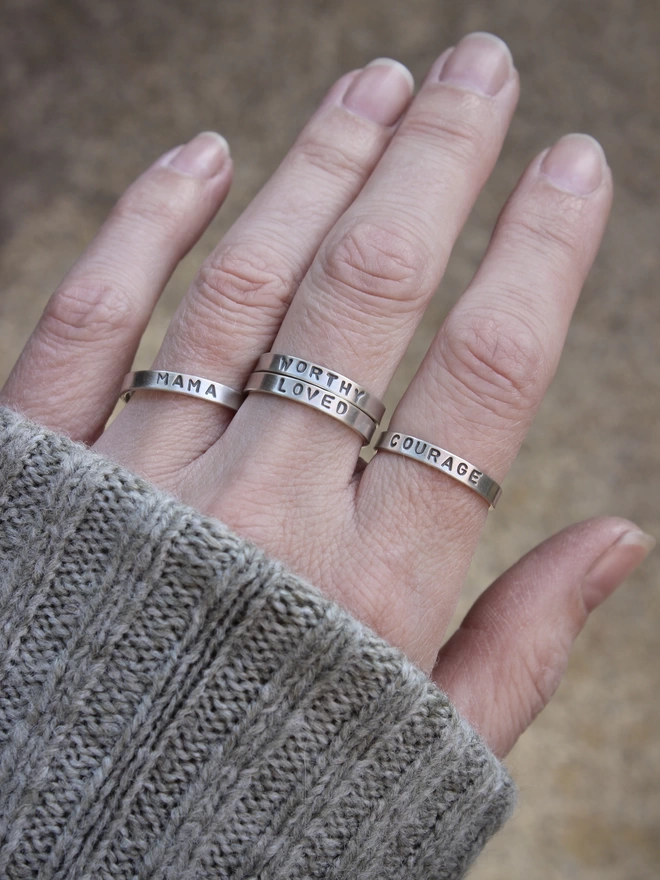 The left hand of a woman in a green knitted jumper, wearing a selection of four simple silver band rings stamped with a single word of empowerment.