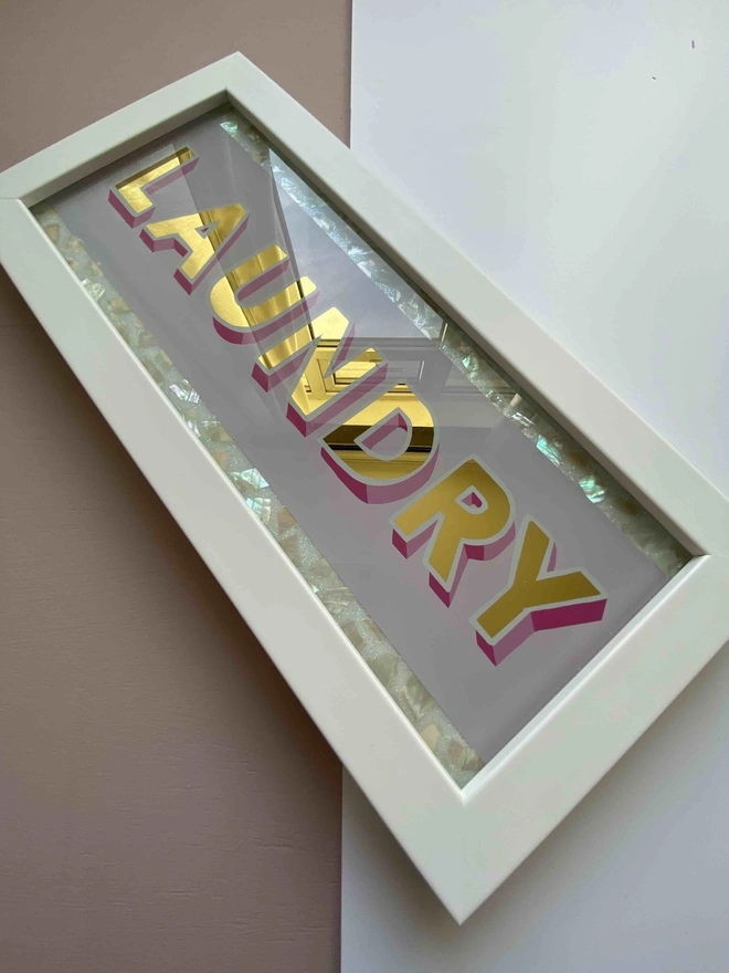 'Laundry' Glass And Gold Leaf Sign, a gold and glass sign sitting on a plain surface in a white frame. 