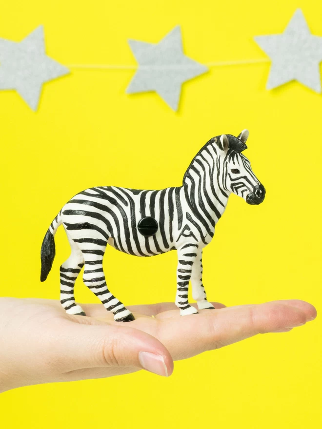  A 9 cm tall black and white striped zebra door handle made of solid plastic stood on the palm of an open hand to show the scale. The zebra has a black bolt head in the middle and is set against a bright yellow background with silver star garlands. The zebra is made of plastic. The brand is Candy Queen Designs.
