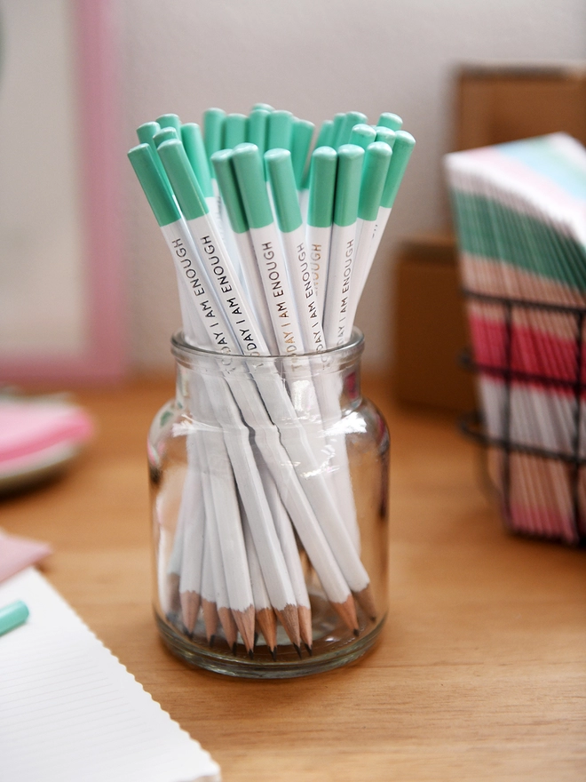 A glass jar full of white pencils with green tips stands on a wooden desk surrounded by various stationery items.