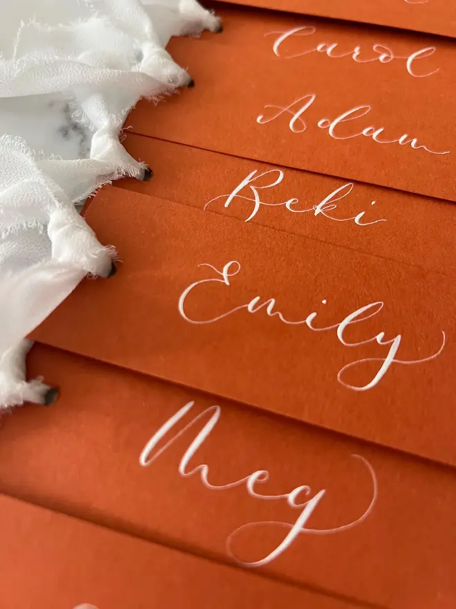 Personalised Calligraphy Rust Orange Place Card, orange place cards with white chiffon ribbons grouped together on a white surface. 