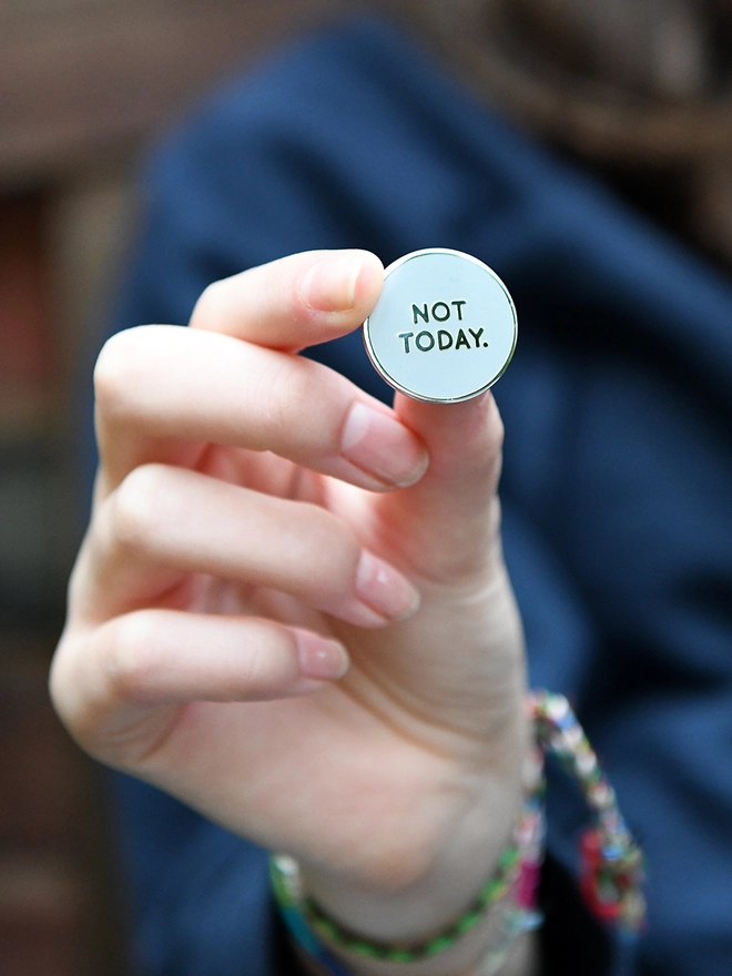 A teenage girl holds a round grey enamel pin badge in her hand that reads "Not Today".