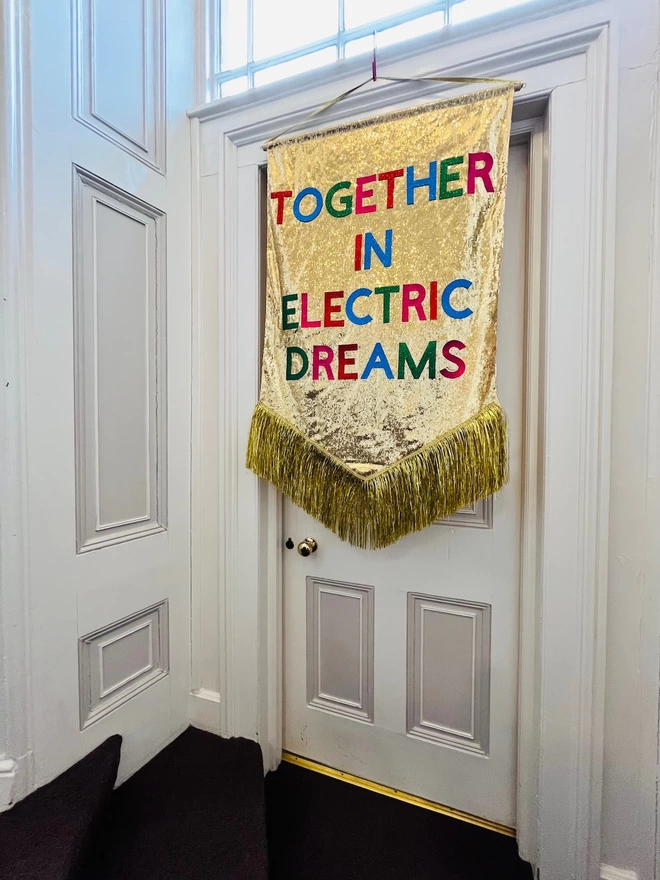 A maxi customisable banner hangs from a hook. It has a gold sequin background and a gold tinsel trim along the bottom. The text is multicoloured and says 'TOGETHER IN ELECTRIC DREAMS'