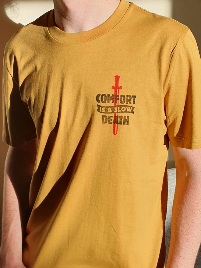 A man wearing  a yellow t-shirt with a black handprinted design on the front depicting a dagger and the phrase 'comfort is a slow death'