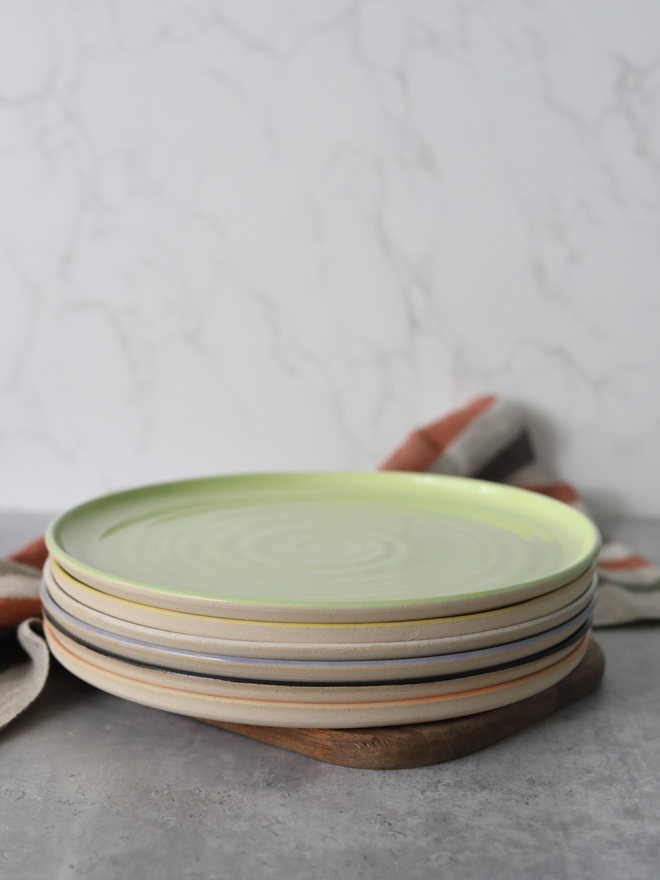 A stack of six handmade ceramic dinner plates in assorted colours — including lime green, yellow, lavender, charcoal, and peach — stacked on a wooden board with a linen napkin draped alongside, set against a white marble background