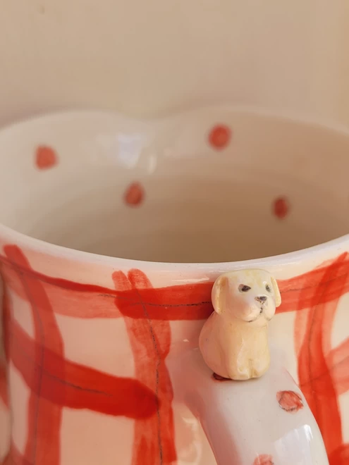 a tiny golden Labrador attached on the handle   of a red gingham check pottery cup