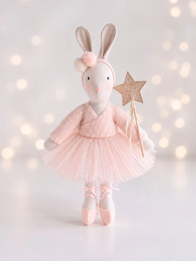 Verity ballerina bunny standing with a gold star wand against a soft pale background with glowing bokeh lights.