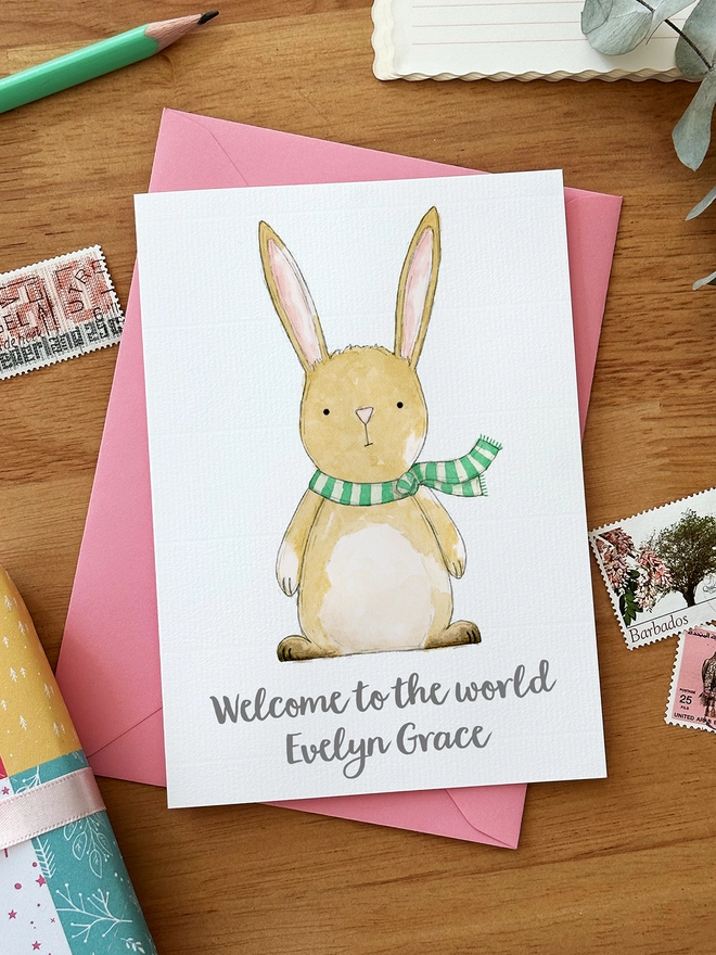 A personalised new baby greetings card with a rabbit illustration lays on a pink envelope on a wooden desk beside various stationery items.