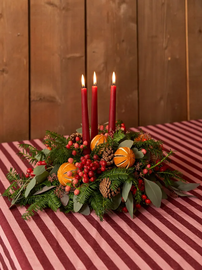 green wreath with dried oranges, red berries, and leaves on a table with red striped table cloth. three lit, red candles in the middle