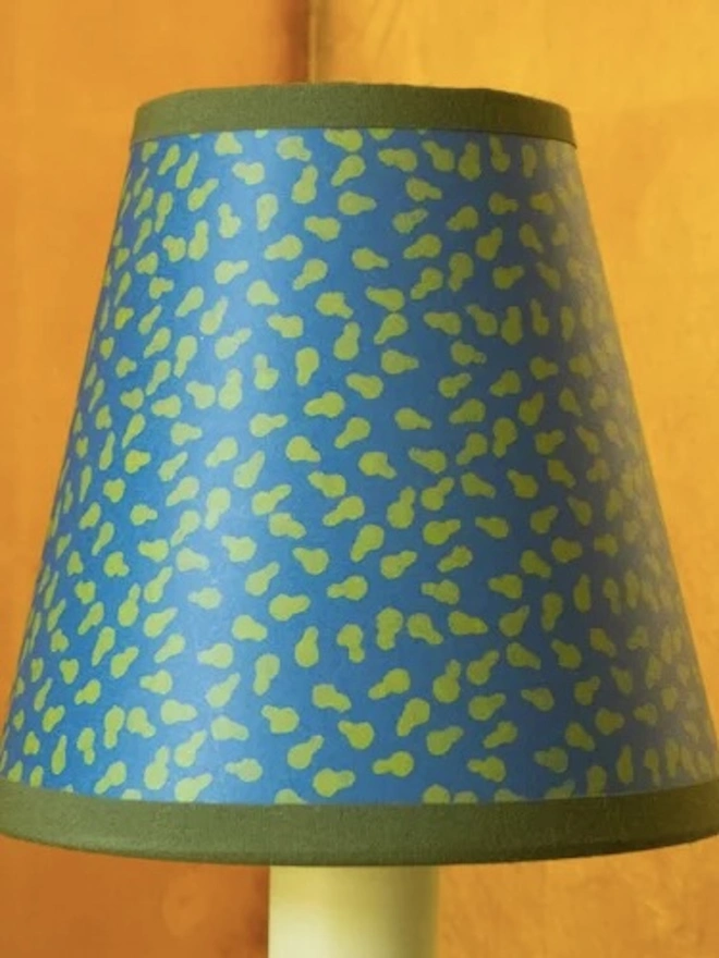 Candle Clip Shades. A small candle lampshade in bright blue with a scattered green leaf print and a deep green trim, set against a warm ochre wall. The bold hand-printed shade adds a playful pop of colour to a wall light or candlestick lamp.