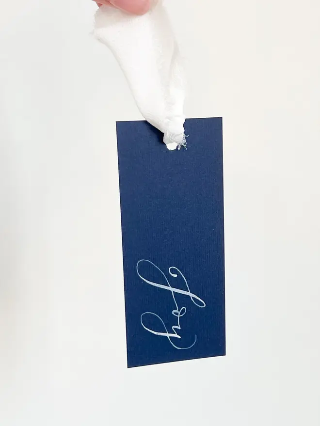 Personalised Calligraphy Wedding Navy Place Cards, navy place card hanging from a hand with a white ribbon. 
