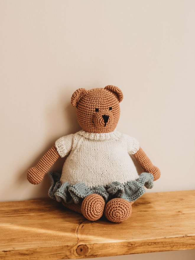 hand crochet brown teddy bear with hand knit white and blue dress on wooden shelf