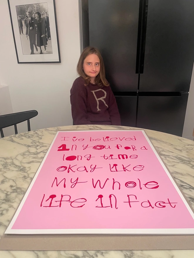 Pink background with red text in Pipers unique font on a large screen print on a marble table with the artist Piper sat proudly behind it 