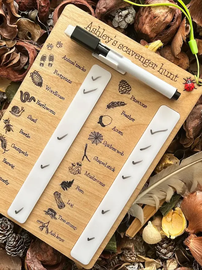 Wooden scavenger hunt collection board engraved with illustrated nature items such as pinecone, feather, acorn, insects, and animal tracks, featuring check-off sliders and a dry-erase pen, displayed on dried leaves and pinecones.