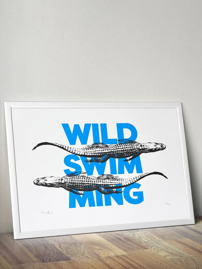 A humorous limited edition A3 screen printed poster. Featuring black alligators over blue 'wild swimming' typography. Mock up in a frame.