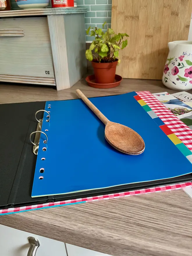 Red gingham recipe binder open on wooden kitchen bench showing 10 colourful dividers.