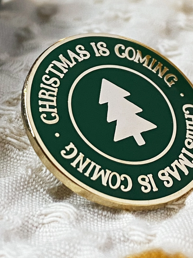 A deep green round enamel pin badge with a gold Christmas tree in the centre and gold lettering around the outside that reads "Christmas Is Coming" rests an ivory fabric.