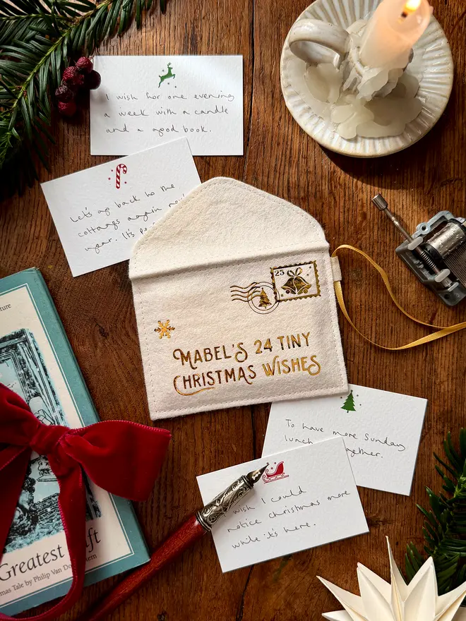 A handmade felt envelope advent calendar with golden lettering that reads 'Mabel's 24 Tiny Christmas Wishes' with several Christmas notecards all laying on a wooden table.