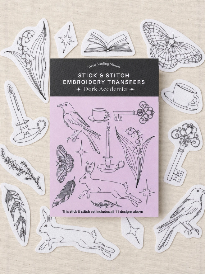 Dark Academia Stick & Stitch embroidery transfers arranged around the product packaging on natural cotton fabric. The set includes 11 water-soluble embroidery designs inspired by gothic literature and nature, including a raven, hare, luna moth, lily of the valley, antique keys, candles, teacups, feathers, stars and an open book. Plastic-free embroidery transfer set by Tired Starling Studio for decorating clothing, accessories and home textiles.